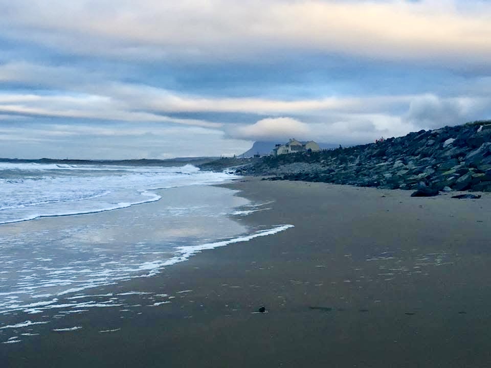 Waves washing onto a quiet sandy beach beside a rocky shoreline under layered blue and cream clouds